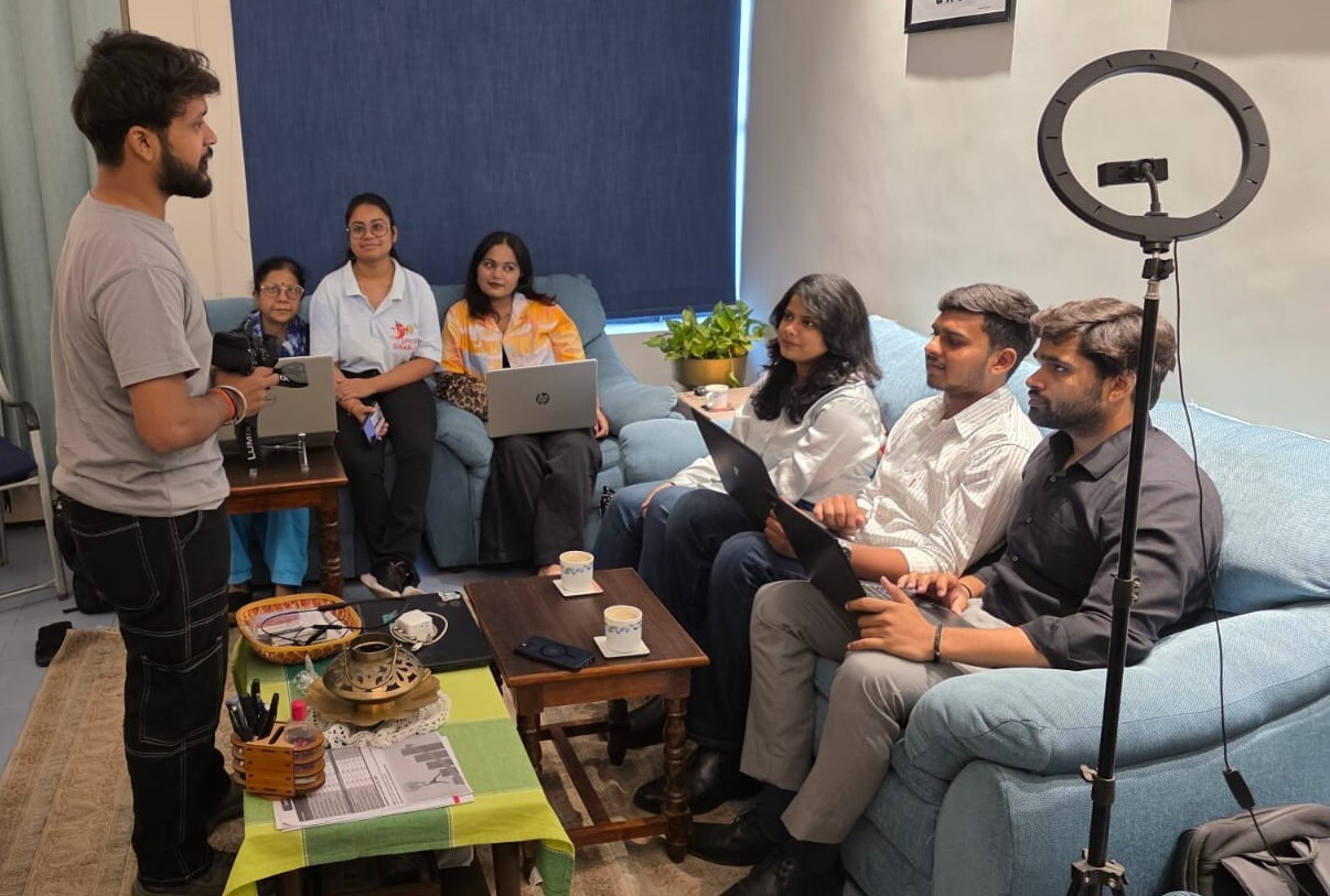 A man stands and speaks to five seated people with laptops in a living room setting, while a ring light and coffee cups are visible on the tables nearby.